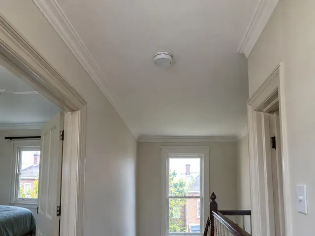 Ceiling-mounted smoke alarm in a painted hallway with doorways and a stair landing in Lewisham